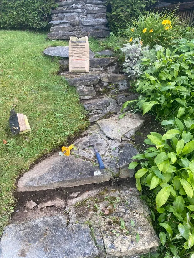 Le haut du chemin, entre le gazon et les plantes : pierres disjointes, sac de mortier et marteau posés sur les marches