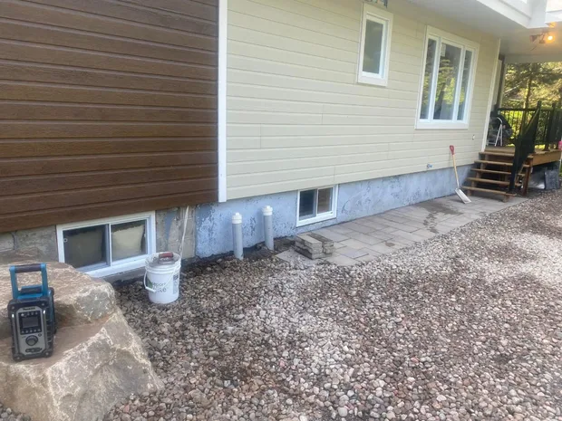 Fondation de béton nu et tachée sous un revêtement jaune pâle, deux tuyaux blancs qui sortent du lit de galets