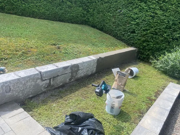 Vieux muret de blocs de pierre grise abîmé le long d'une haie de cèdres, chaudières et sac de ciment posés sur le gazon