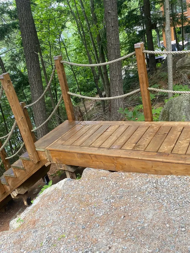 Grand palier de bois teint fraîchement fini, cordes de rampe posées, gros bloc de roc et gravier au pied