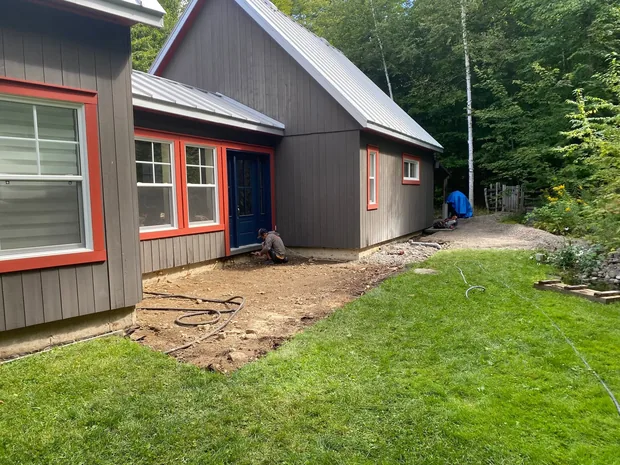 Devant une maison grise à cadres orange et porte bleue, le sol en terre battue où le patio va se bâtir, Joe accroupi au pied du mur