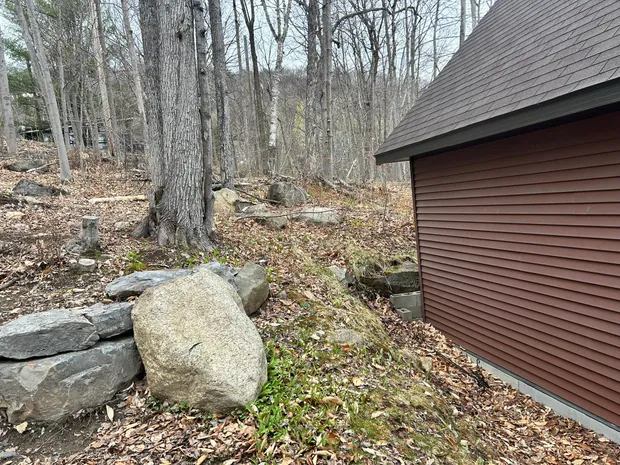 Le pied d'un bâtiment brun-rouge disparaît sous les feuilles mortes, un gros bloc de pierre pâle à côté